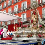 Procesión de la Almudena en Madrid. Una imagen de la Virgen con el Niño sobre un trono adornado con flores, con el fondo de edificios rojos y gente vestida de gala.