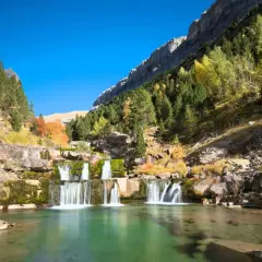 Cascada en un valle montañoso, ideal para rutas senderismo España. El agua verde turquesa fluye entre rocas y árboles otoñales.