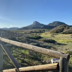 Un sendero de madera bordea un paisaje verde y montañoso, con el imponente monte de las cenizas al fondo bajo un cielo azul.
