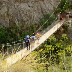 Senderismo en las **rutas Comunidad Valenciana**. Personas cruzan un puente colgante entre riscos y vegetación.
