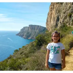 Niña sonriente en la ruta acantilados Benitachell, con el mar azul y formaciones rocosas al fondo.