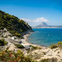 La cala del francés, con sus casitas blancas y el mar azul, se asoma a un paisaje rocoso y verde. Al fondo, la silueta de una montaña domina la costa.