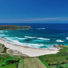 Vista aérea de la playa de Pantín con olas turquesas rompiendo en la arena dorada, rodeada de verdes colinas y el azul intenso del océano.