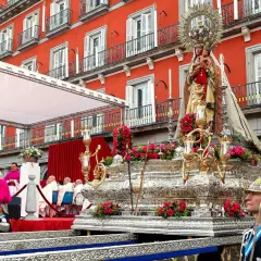 Procesión de la Almudena en Madrid. Una imagen de la Virgen con el Niño sobre un trono adornado con flores, con el fondo de edificios rojos y gente vestida de gala.