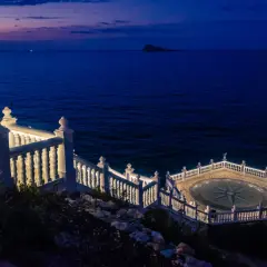 El mirador de Benidorm de noche, con el mar azul oscuro y un cielo crepuscular. Una balaustrada blanca ilumina el camino hacia una plataforma circular con una rosa de los vientos.
