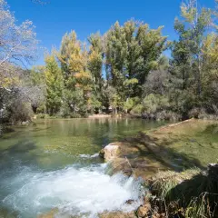 Un río de aguas cristalinas con un pequeño salto de agua. Al fondo, árboles con hojas otoñales y un cielo azul. ¡Un lugar con **alto tajo que ver**!