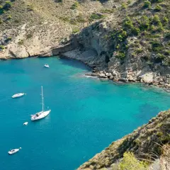 La cala del metge, con aguas turquesas y barcos navegando, se abre paso entre acantilados rocosos y vegetación mediterránea.