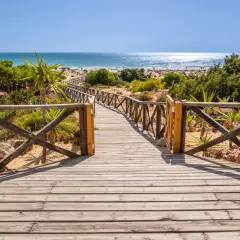 Pasarela de madera hacia la playa en Sancti Petri. El paseo marítimo se abre a un mar azul brillante y arena dorada.