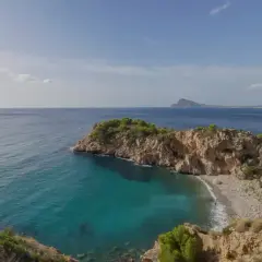 Vista de una cala con aguas turquesas y una gran barra de guijarros, rodeada de acantilados rocosos y vegetación.