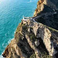 Faro blanco sobre acantilado rocoso en el **parque natural de la Sierra Helada**, con mar turquesa y camino serpenteante.