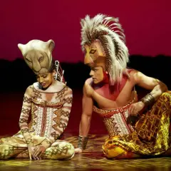 Dos actores con trajes de león, uno con máscara de leona y otro con melena, sentados en el escenario. La duración del Rey León en Madrid es inolvidable.