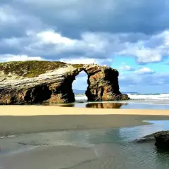 Arco natural en una playa de arena dorada, con el mar y nubes dramáticas. Un ejemplo de las mejores playas Galicia.