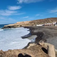 Olas rompen en la playa de arena negra de los charcones de Ajuy, con casas blancas al fondo y acantilados dorados.