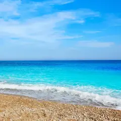 Aguas turquesas bañan la playa de guijarros de cala dels testos, con un acantilado rocoso a la izquierda y un cielo azul despejado.