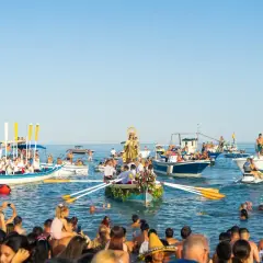 Procesión marítima con la Virgen del Carmen en un barco, rodeada de embarcaciones y gente en el agua, celebrando las fiestas.