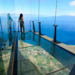 Mujer en mirador de cristal, 3 metros sobre el cielo, con vistas al mar y un pueblo.