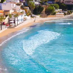 La playa de cala el Portet, con sus aguas turquesas y arena dorada, se extiende bajo un cielo azul, flanqueada por palmeras y casas blancas.