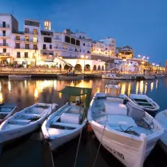 Noche en el paseo marítimo de L'Ametlla de Mar. Barcas blancas flotan en el puerto, reflejando las luces de los edificios y restaurantes.