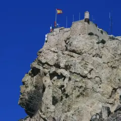 La cara del moro alicante, una roca con forma de perfil humano, corona la cima de una montaña rocosa con un castillo y una bandera española.