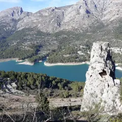 Impresionante vista del embalse de Guadalest con su agua turquesa, rodeado de montañas y pinos. Una roca solitaria se alza como centinela.