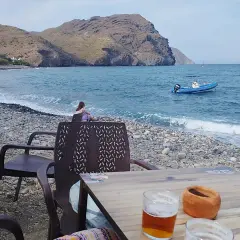 Una persona contempla el mar en la playa de las negras opiniones. Un bote azul descansa en el agua, mientras las olas acarician la costa pedregosa.