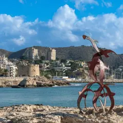 Escultura de aves sobre rocas, con el pueblo de Moraira y su torre defensiva al fondo.