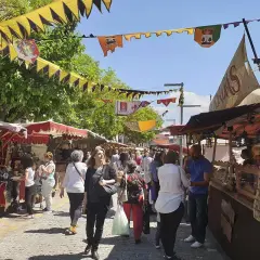 Gente pasea por el mercadillo medieval El Álamo, con puestos de artesanía y comida bajo banderines coloridos.