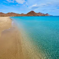 Arena dorada y aguas cristalinas en una de las playas bonitas en España, con montañas áridas al fondo bajo un cielo azul.