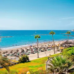 Un día soleado en el paseo marítimo de Playa de las Américas, con gente disfrutando de la playa, sombrillas y palmeras.