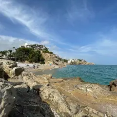 Rocas doradas en primer plano, la caleta Villajoyosa con gente disfrutando del mar azul y edificios blancos en la colina.