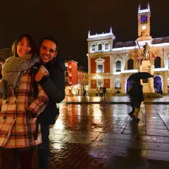Pareja sonriente en la Plaza Mayor de Valladolid, un lugar bonito que ver en Valladolid, con edificios iluminados y el suelo mojado por la lluvia.
