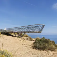 El mirador del faro de Santa Pola se proyecta sobre el mar azul, con vegetación árida y un cielo despejado.