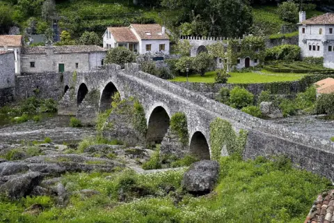 Puente de piedra cubierto de hiedra, con casas tradicionales al fondo. Un lugar idílico para visitar, así es como ponte Maceira como llegar.