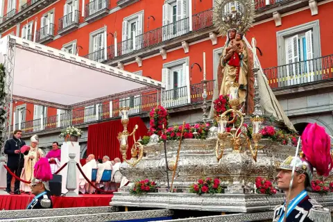 Procesión de la Almudena en Madrid. Una imagen de la Virgen con el Niño sobre un trono adornado con flores, con el fondo de edificios rojos y gente vestida de gala.