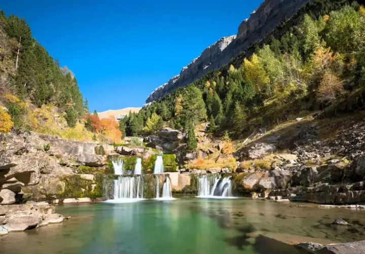Cascada en un valle montañoso, ideal para rutas senderismo España. El agua verde turquesa fluye entre rocas y árboles otoñales.