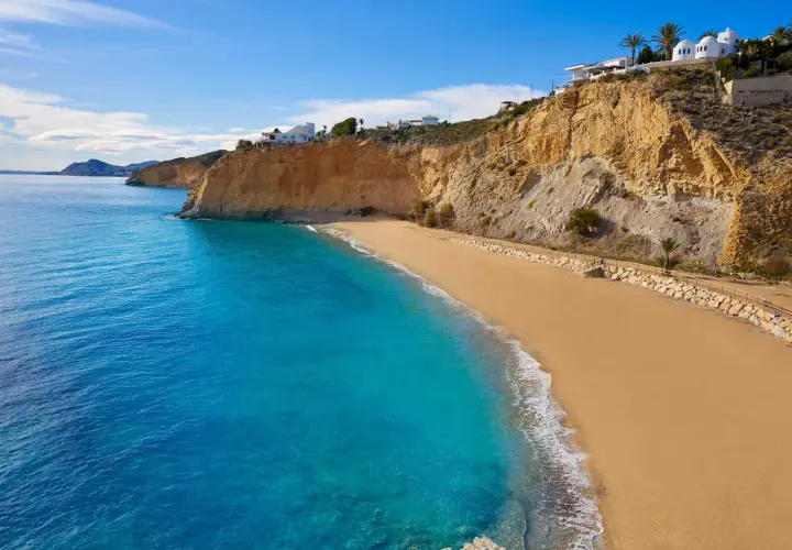 Arena dorada y aguas turquesas en la playa de Villajoyosa, con acantilados y casas blancas al fondo.