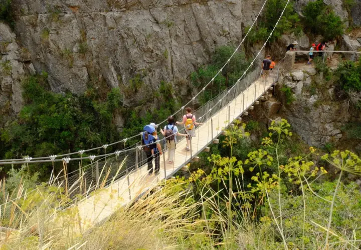 Senderismo en las **rutas Comunidad Valenciana**. Personas cruzan un puente colgante entre riscos y vegetación.