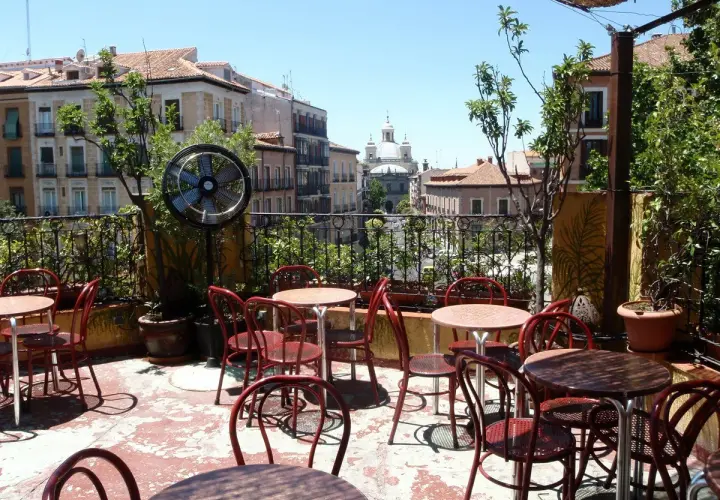 Terraza de verano en Madrid, con mesas y sillas rojas, ventilador y vistas a edificios históricos. Un lugar perfecto para el viajero madrileño.