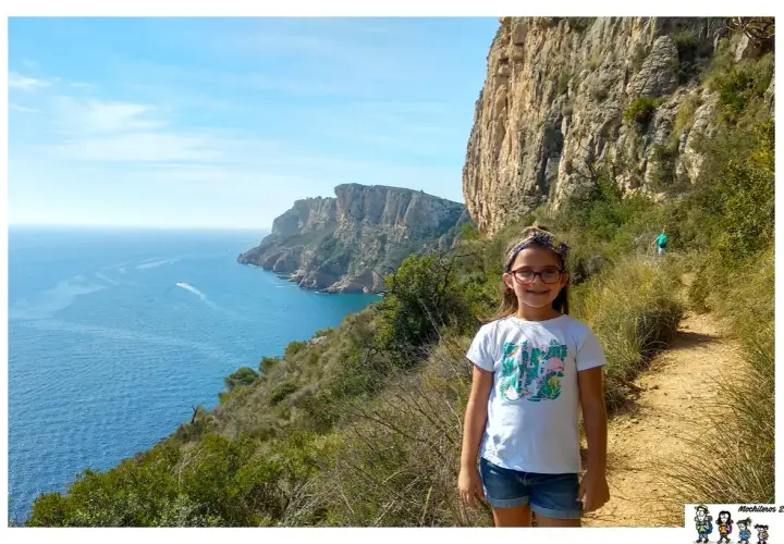 Niña sonriente en la ruta acantilados Benitachell, con el mar azul y formaciones rocosas al fondo.