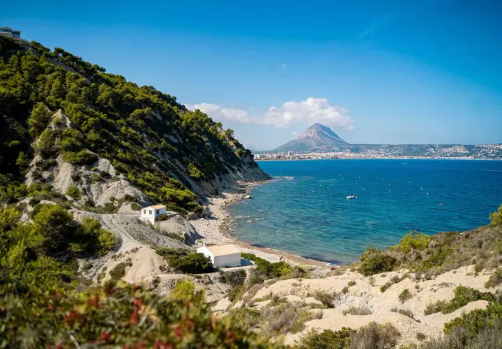 La cala del francés, con sus casitas blancas y el mar azul, se asoma a un paisaje rocoso y verde. Al fondo, la silueta de una montaña domina la costa.