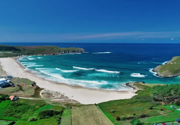 Vista aérea de la playa de Pantín con olas turquesas rompiendo en la arena dorada, rodeada de verdes colinas y el azul intenso del océano.