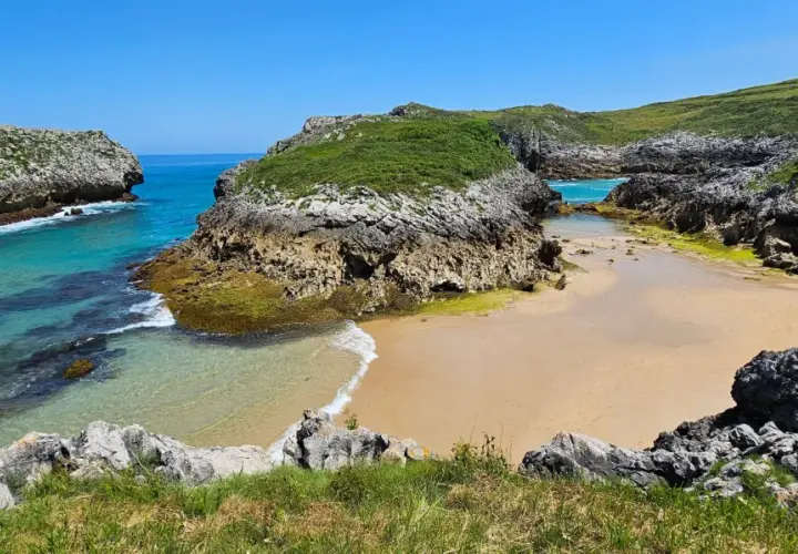 Una cala asturiana con aguas turquesas y arena dorada, rodeada de acantilados verdes. Un paraíso entre las playas más bonitas de Asturias.