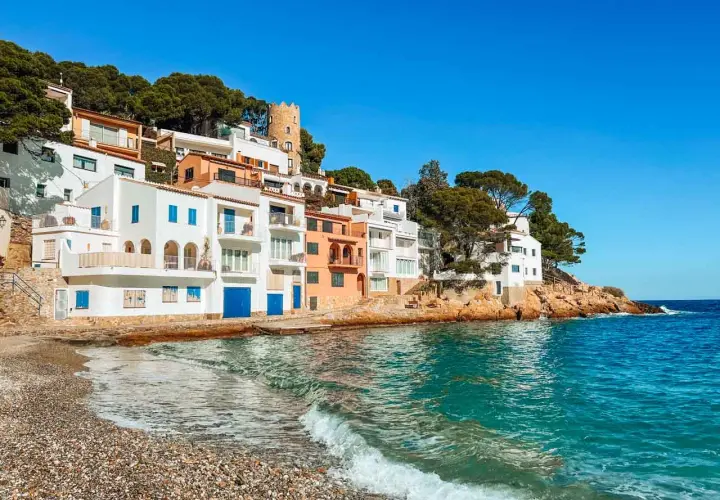 Casas blancas y coloridas se asoman al mar turquesa en la costa brava españa. Una pequeña playa de guijarros recibe las olas.