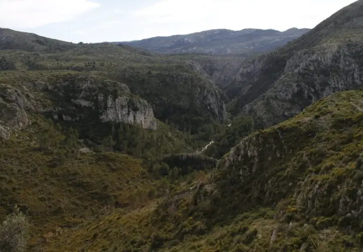 Paisaje montañoso con vegetación verde y rocas escarpadas. El **barranc de l'infern** se abre paso entre las colinas, con un camino serpenteante.