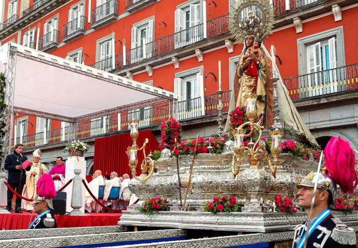Procesión de la Almudena en Madrid. Una imagen de la Virgen con el Niño sobre un trono adornado con flores, con el fondo de edificios rojos y gente vestida de gala.