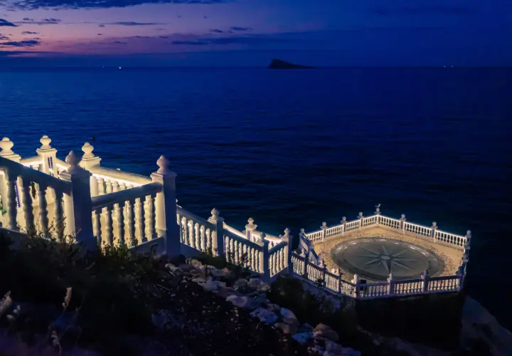 El mirador de Benidorm de noche, con el mar azul oscuro y un cielo crepuscular. Una balaustrada blanca ilumina el camino hacia una plataforma circular con una rosa de los vientos.