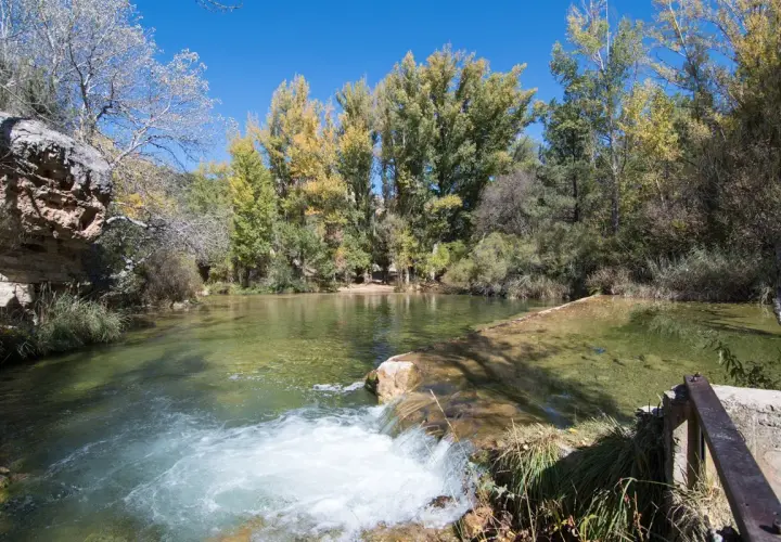 Un río de aguas cristalinas con un pequeño salto de agua. Al fondo, árboles con hojas otoñales y un cielo azul. ¡Un lugar con **alto tajo que ver**!