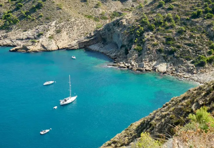 La cala del metge, con aguas turquesas y barcos navegando, se abre paso entre acantilados rocosos y vegetación mediterránea.