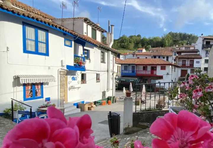 Casas blancas con detalles azules, tejados rojos y flores rosadas en un día soleado. Un vistazo a los encantadores pueblos cerca de Cudillero.