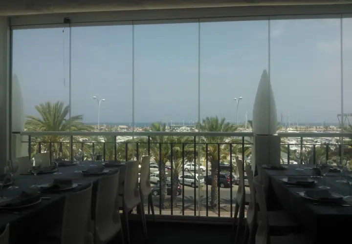 Mesas puestas con vistas al puerto deportivo de la barqueta Denia. Palmeras y barcos crean un ambiente vacacional.